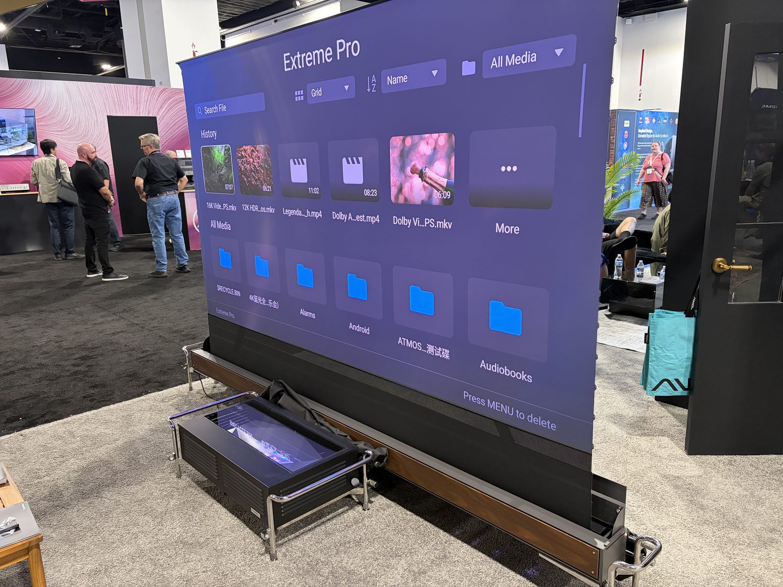 An AWOL Vision motorized screen rising out of a wood-trimmed floor base at CEDIA Expo, with an ultra short throw projector sitting in a vented black enclosure with chrome handles in front of it