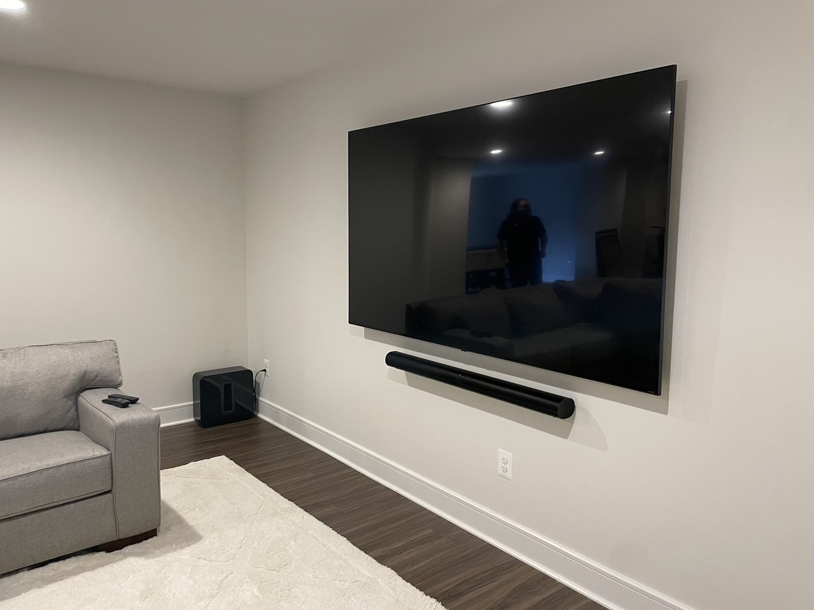 A very large wall-mounted TV with a long soundbar mounted beneath it and a subwoofer on the floor in the corner of a family room