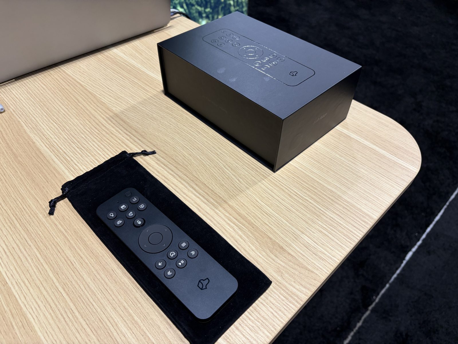 A black Josh Edge handheld remote lying on a velvet pouch next to its retail box, showing a round navigation pad with media buttons below it