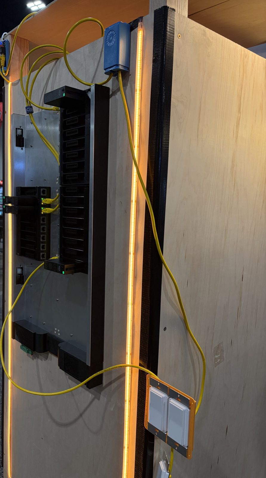 A low-voltage lighting panel on a plywood display wall at CEDIA Expo, with yellow network patch cables, glowing LED strip lighting along the edges, and wall switches below