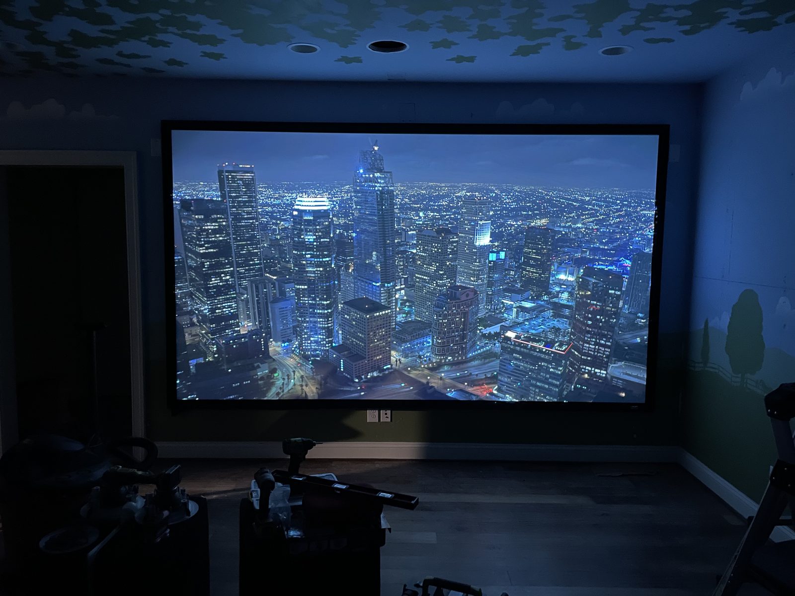 A projector screen showing a city skyline at night on the wall of a playroom with a painted sky and meadow mural, installation tools still on the floor
