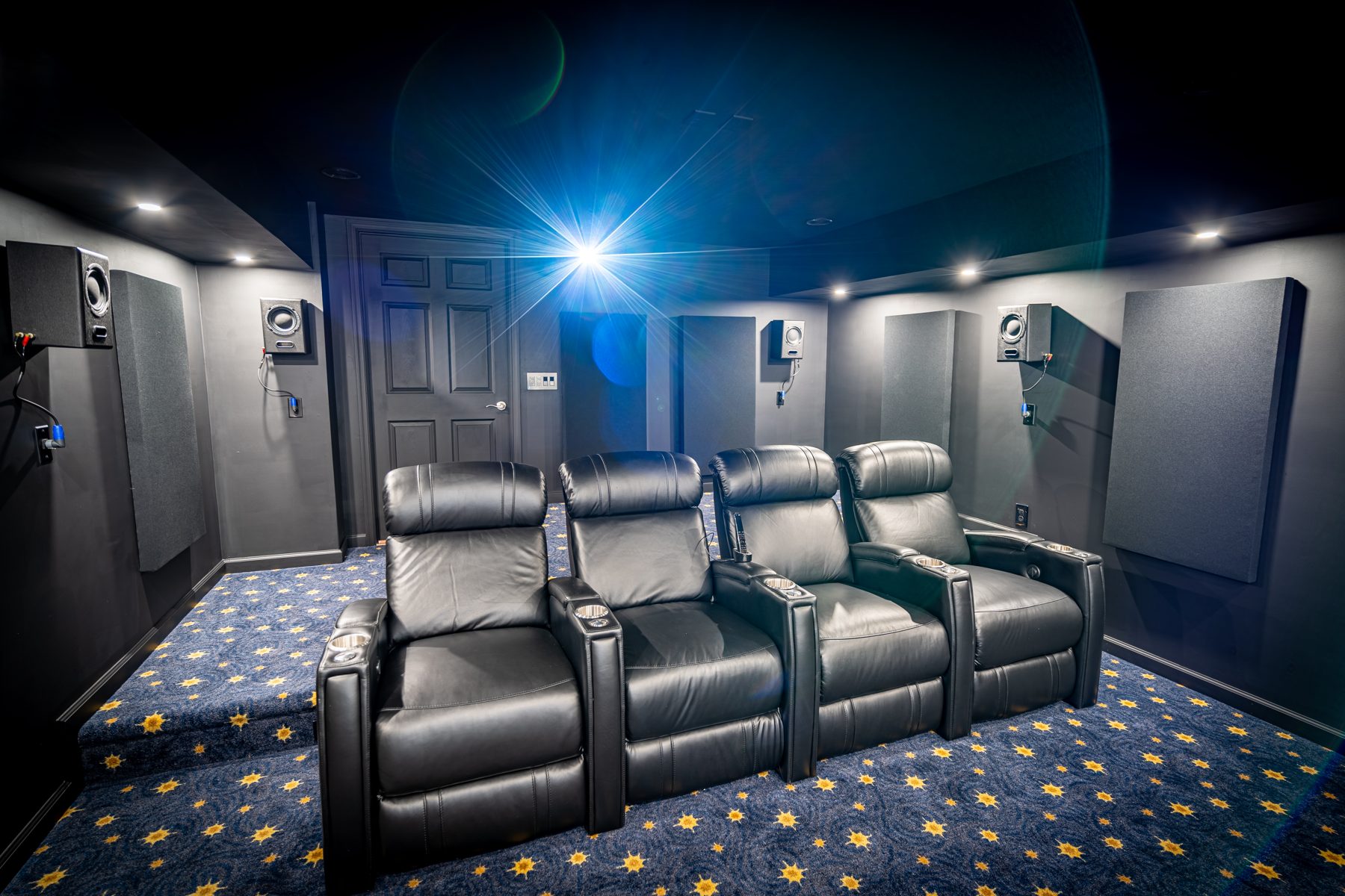 The demo theater seen from the front: four black leather recliners on a star-pattern carpet, wall-mounted surround speakers and acoustic panels either side