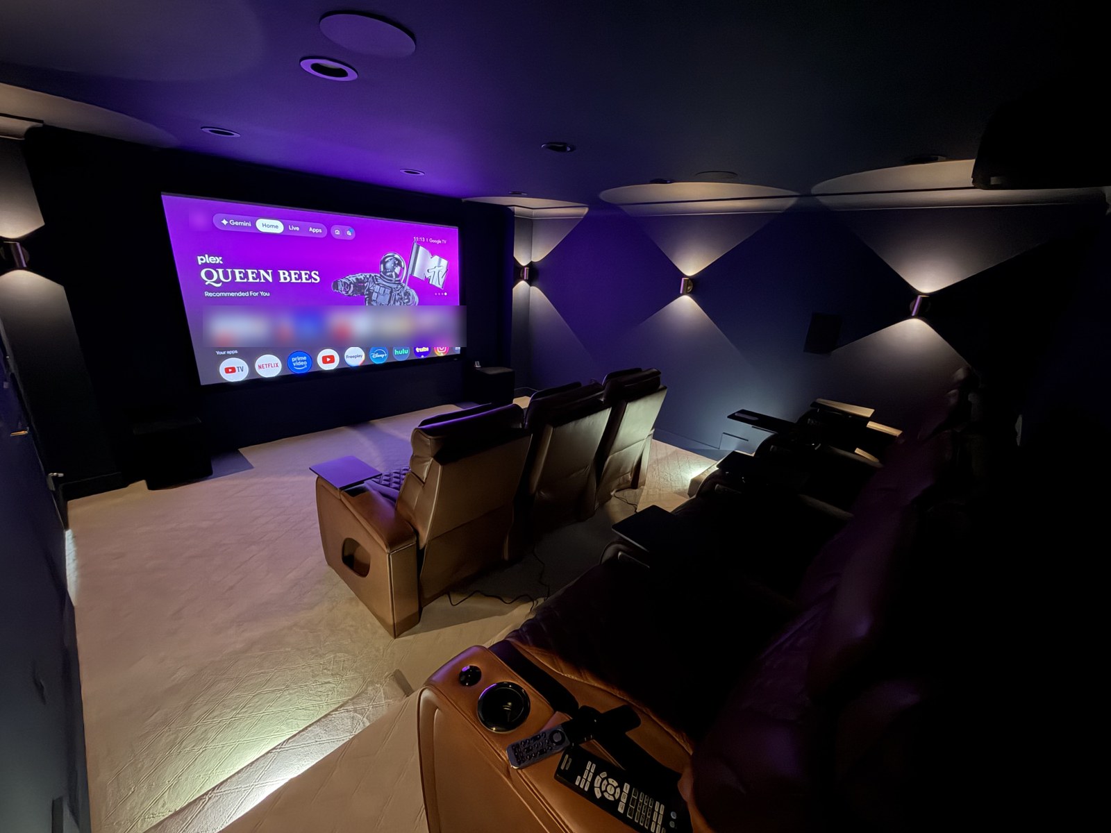 A dark home theater with tan leather recliners facing a large projection screen showing a Google TV home screen; two remotes rest on the arm of the front chair